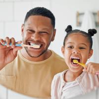A father and his young daughter brushing their teeth together in a bathroom.